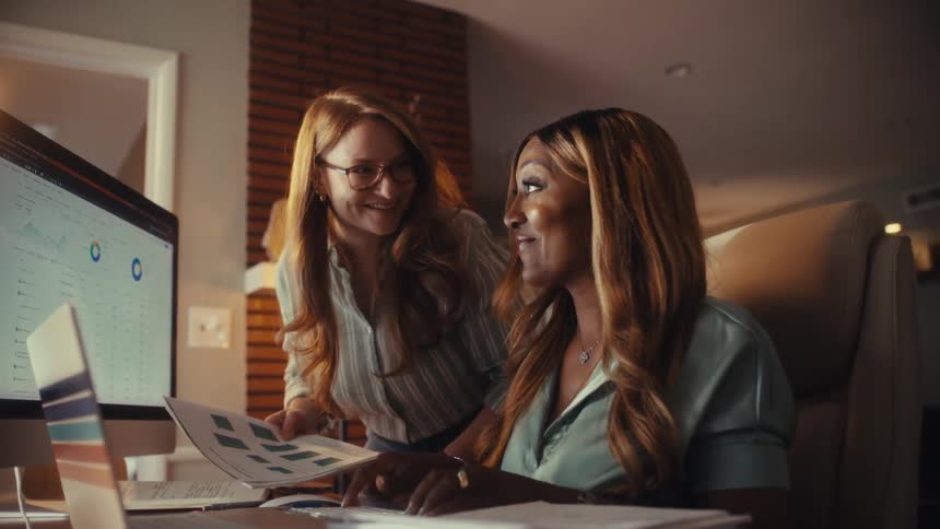 Two diverse businesswomen collaborating in a home office review financial data on their computers and happily celebrate their success