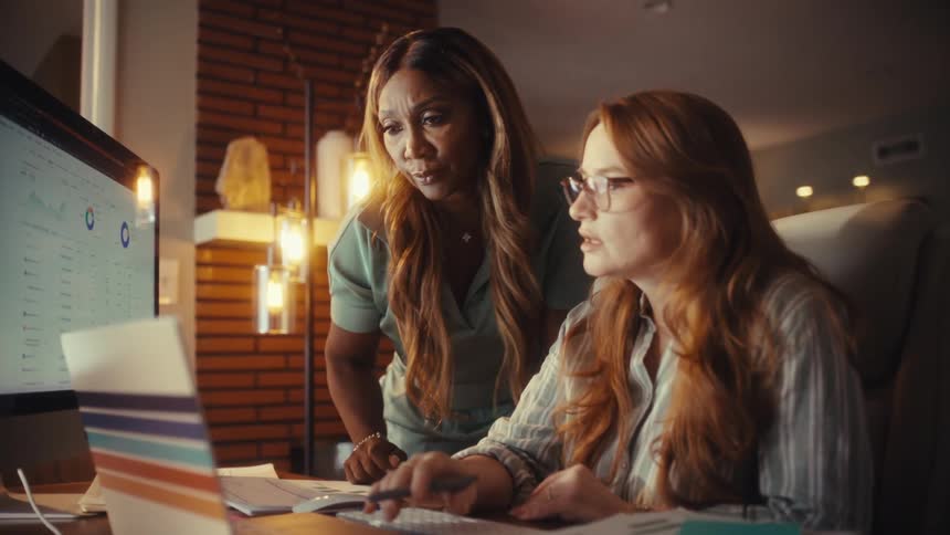 Two focused businesswomen collaborate in a dimly lit office, discussing financial charts and data displayed on a computer monitor
