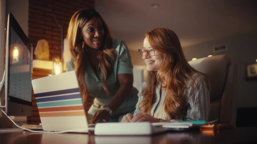 Two diverse female colleagues collaborate on a project in a home office, celebrating a successful outcome with smiles and laughter