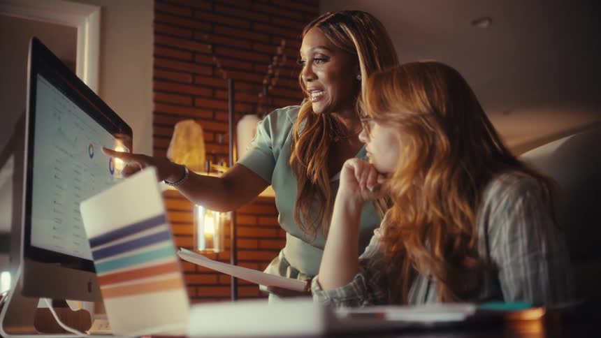Two diverse businesswomen collaborate in a modern office at night, analyzing data and charts on a computer screen