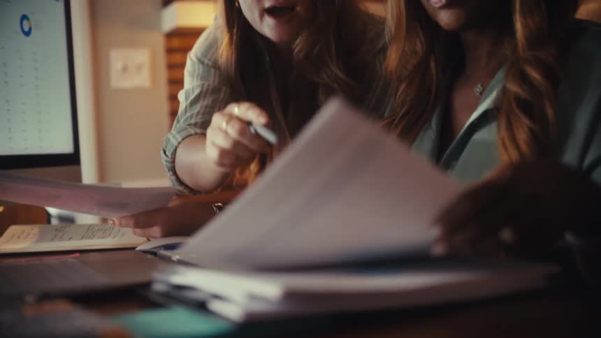 Two diverse female colleagues work together in a warmly lit home office, analyzing data from charts and paperwork