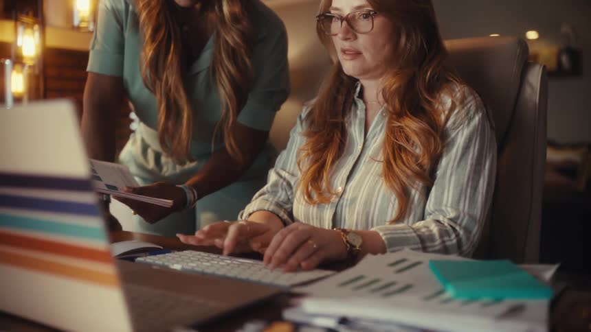 Two focused businesswomen collaborate late into the night in a modern home office, analyzing data on a laptop and reviewing documents