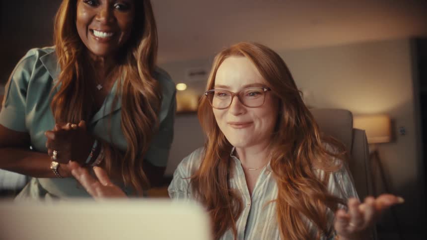 Two diverse women excitedly celebrate a victory while looking at a laptop in a warmly lit room at night