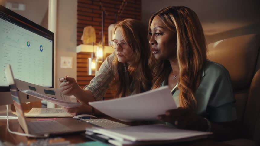 Two diverse businesswomen work together late into the evening, analyzing financial reports and charts on a computer in their office