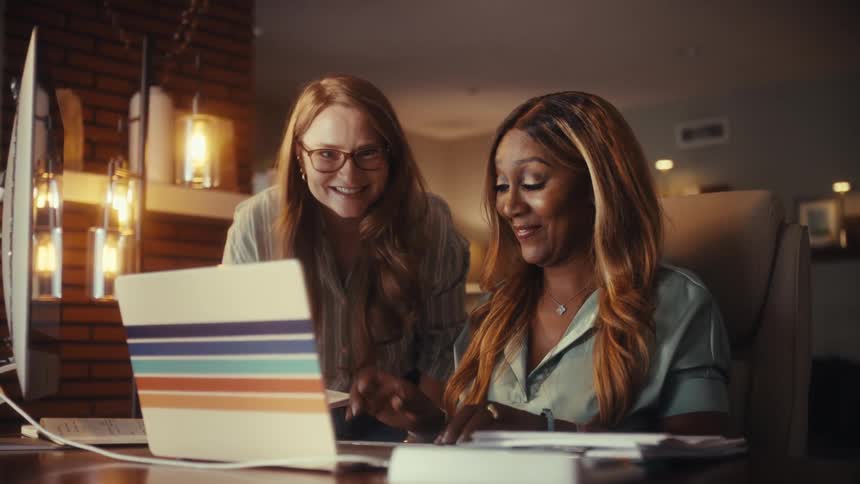 Two diverse businesswomen happily celebrate a major success, laughing and cheering together while working on a laptop in their home office