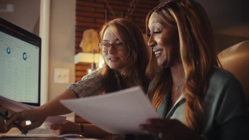 Two diverse female colleagues collaborate positively while analyzing business data and charts on a computer in a modern office