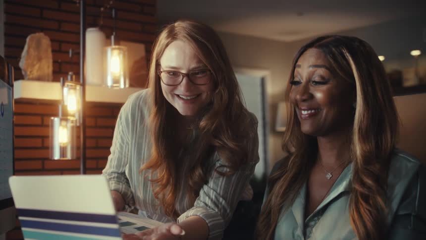 Two diverse female colleagues share a moment of joy and success as they review positive results on their computers in a stylish office