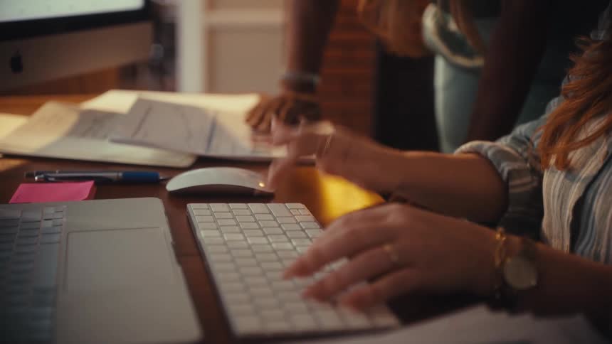 A close-up slow-motion shot of a woman's hands typing on a keyboard while a colleague looks on in the background