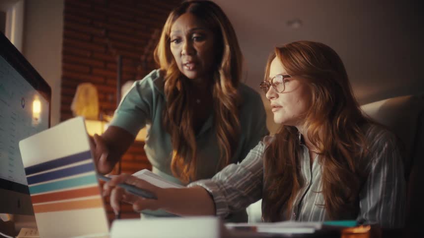 Two diverse professional women collaborate late at night in a warmly lit office, analyzing data on a computer screen