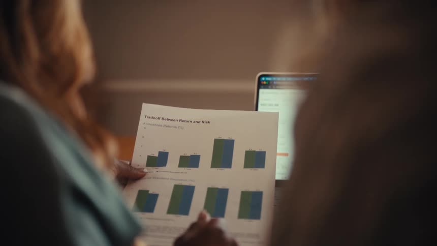 Over-the-shoulder view of two colleagues collaborating and analyzing financial charts on a laptop and a printed report