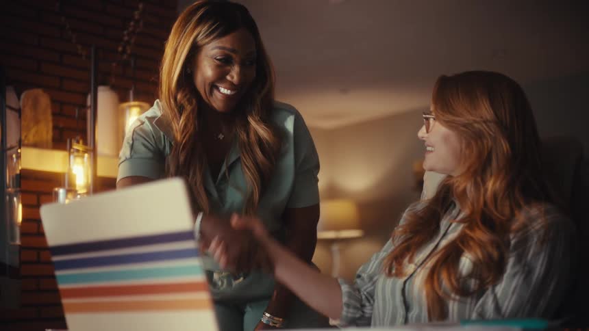 Two diverse businesswomen smile and shake hands in agreement after a successful collaboration on a laptop in their office at night