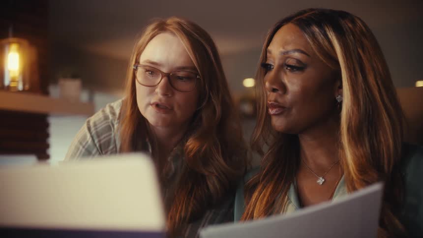 Two diverse businesswomen collaborate on a project, excitedly discussing ideas while pointing at a laptop screen in a warmly lit office