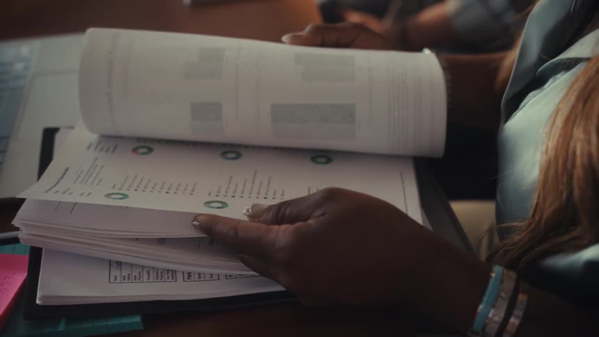 A woman's hands slowly flip through pages of a business report containing charts and data at an office desk