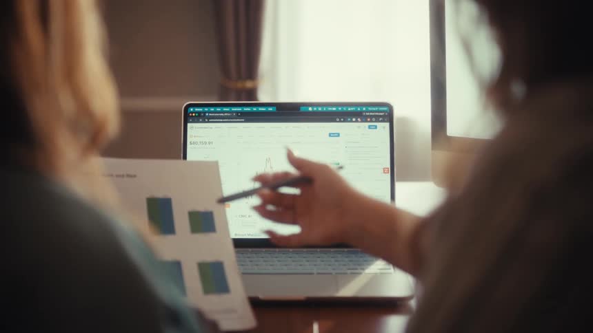 Over the shoulder shot of two business professionals analyzing financial charts and graphs on a laptop and paper documents