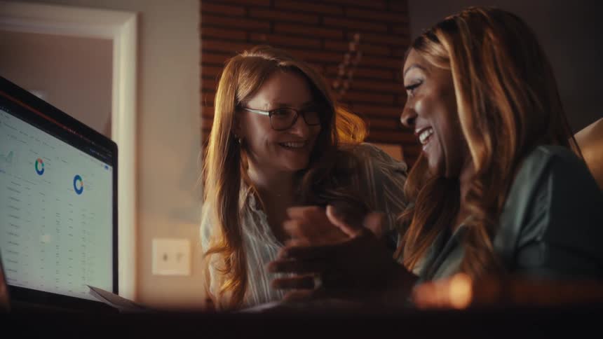 Two diverse female colleagues analyze data charts on a computer, celebrate their success with smiles and a happy embrace in their office