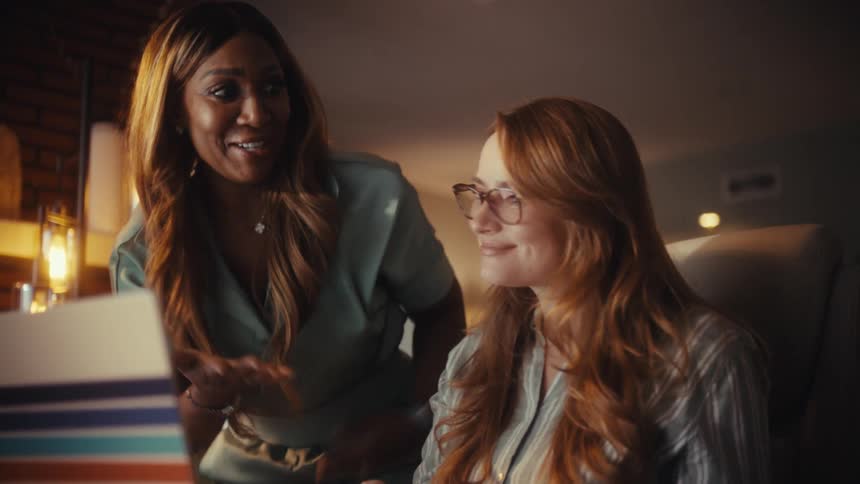 Two diverse female coworkers happily celebrate a successful work project on their laptop in a warmly lit office at night