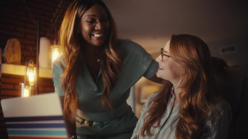 Two diverse businesswomen collaborate on a laptop in a cozy office at night, smiling and hugging in celebration of their successful teamwork