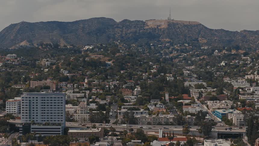 A sweeping high-angle aerial view of the Los Angeles cityscape with the iconic Hollywood sign on the distant hills and traffic moving on the freeway below