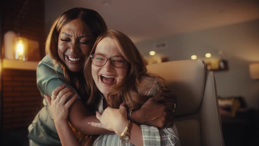 Two diverse female entrepreneurs celebrate a success on their laptop, laughing and hugging joyfully in a cozy home office at night