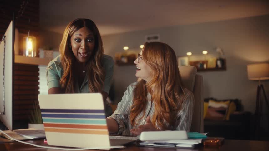 Two diverse female entrepreneurs joyfully celebrate a business victory while working late in their home office