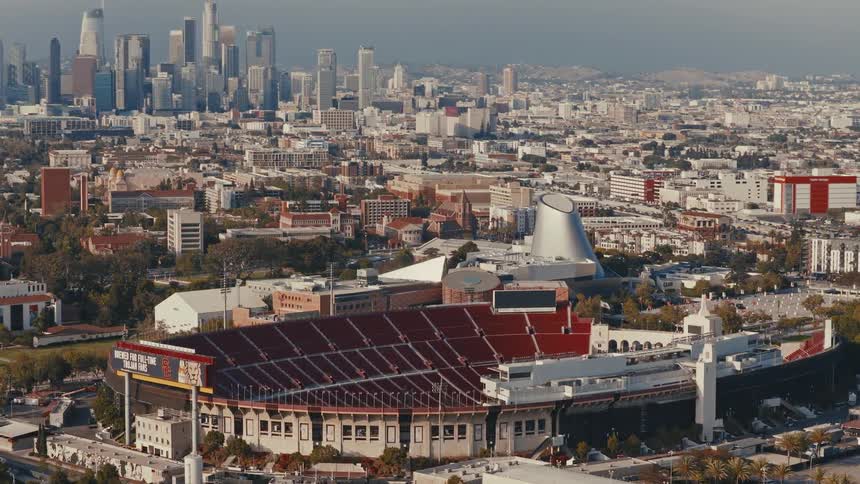 A static telephoto shot of the LA Memorial Coliseum with the downtown LA skyline in the background and a bright sun glare reflecting off a campus building