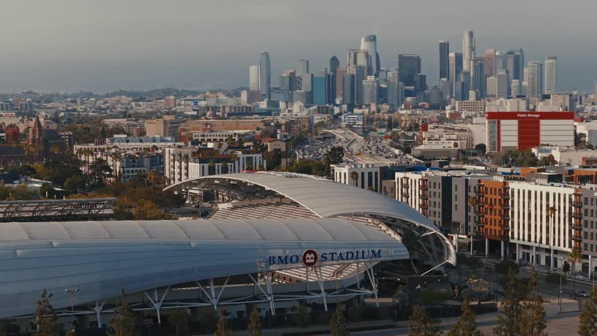A wide aerial shot of BMO Stadium with the downtown Los Angeles skyline and a busy freeway in the background