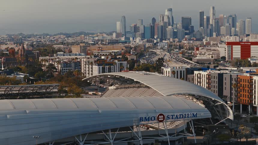 A sweeping aerial view reveals the iconic BMO Stadium set against the sprawling Los Angeles cityscape and a busy freeway
