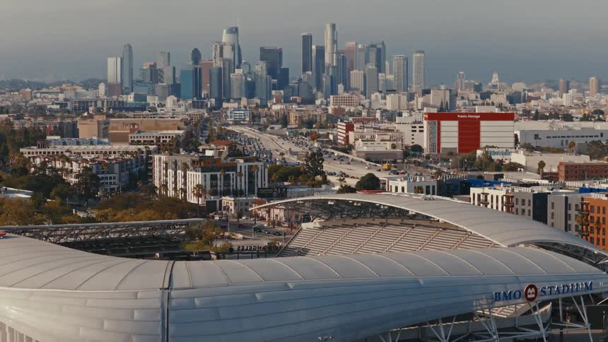 A cinematic aerial view pans across BMO Stadium with the downtown Los Angeles skyline and a busy freeway in the background