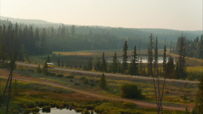 A yellow camper van drives on a scenic road through a hazy evergreen forest and past a tranquil lake