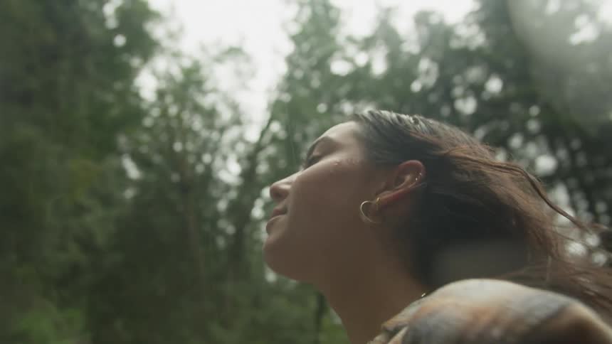 A young woman in a plaid shirt smiles peacefully with her eyes closed as she feels the mist from a powerful waterfall in a lush forest