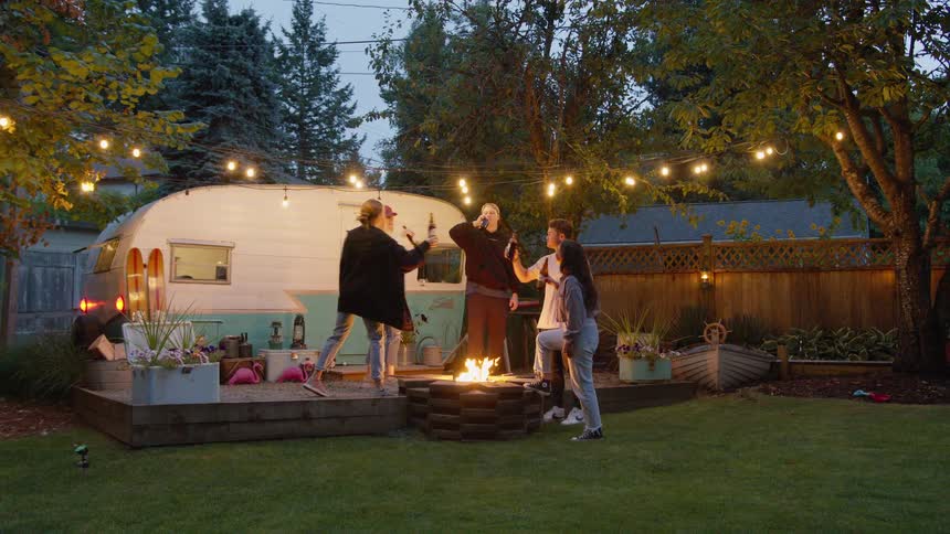 A diverse group of young friends celebrate with a toast in slow motion around a fire pit during a backyard party next to a retro camper