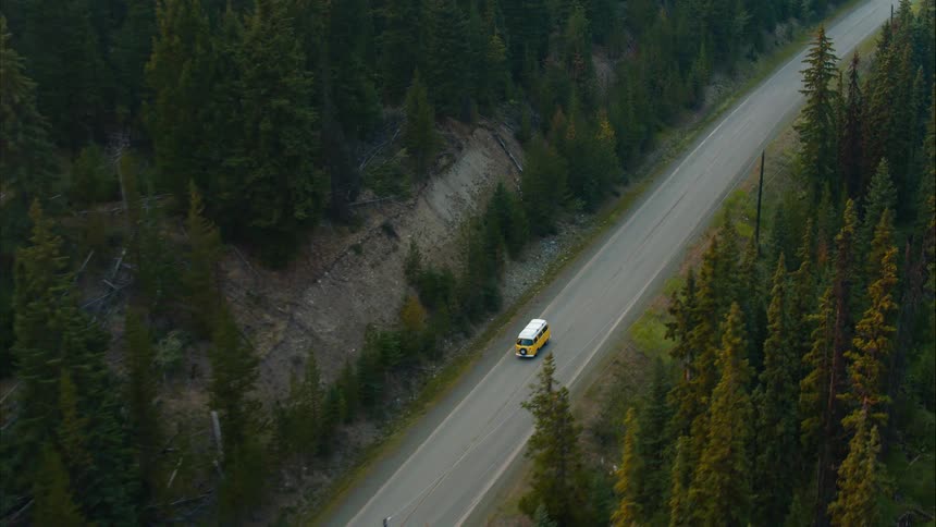 High angle aerial view of a yellow vintage van driving on a scenic mountain road surrounded by a lush green pine forest