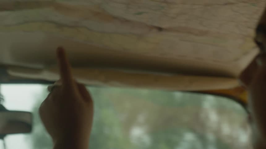 A young woman traces a route on a map taped to the roof of a vintage car during a road trip