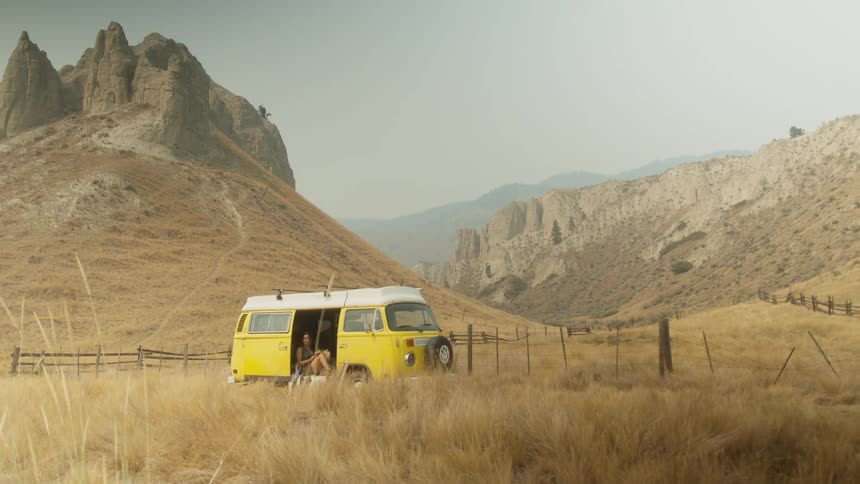 A couple relaxes in the open door of their yellow vintage camper van, parked in a scenic and arid mountain valley during a road trip