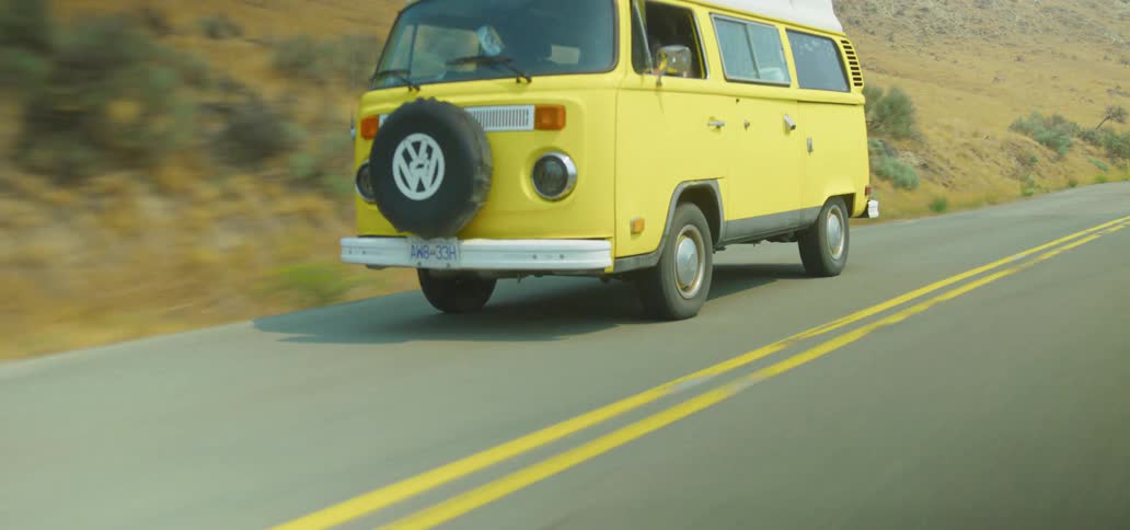 A classic yellow Volkswagen camper van cruises down a scenic country road through dry, rolling hills