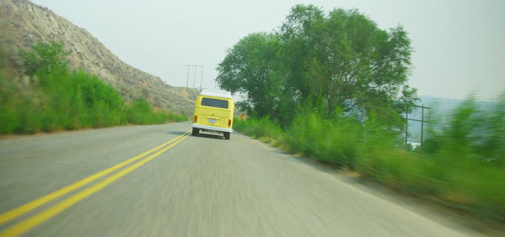 A point-of-view shot follows a classic yellow Volkswagen camper van on a road trip down a winding country road