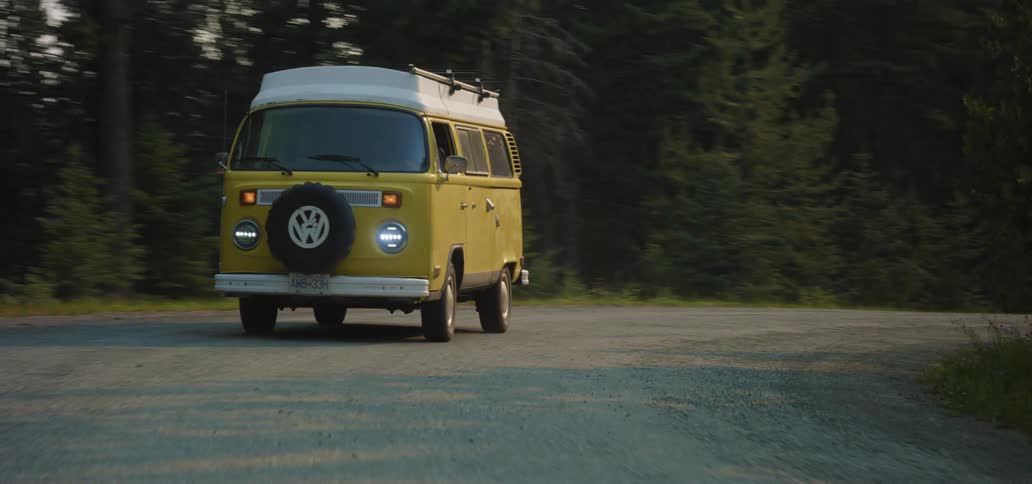 A classic yellow Volkswagen Westfalia camper van drives towards the camera on a winding mountain road through a dense forest at dusk
