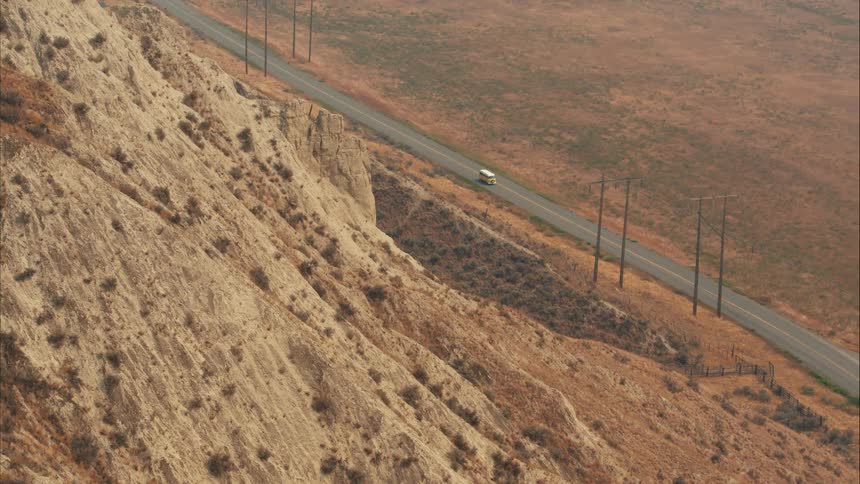 An aerial view shows a lone vehicle driving on a remote highway alongside a steep, eroded bluff in a vast, arid landscape
