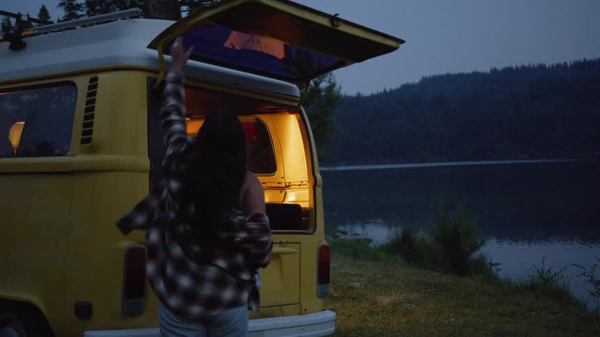 A solo female traveler enjoys a quiet moment by the lake at dusk, using her phone before closing up her vintage yellow camper van for the night