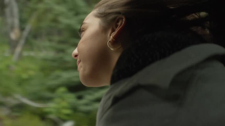 A carefree woman enjoys the fresh air with her head out of a moving car window as she drives through a lush green forest