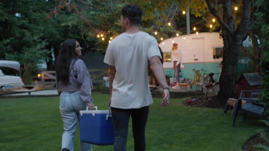 A young couple carries a blue cooler as they walk across a lawn to join their friends at a summer backyard party in slow motion