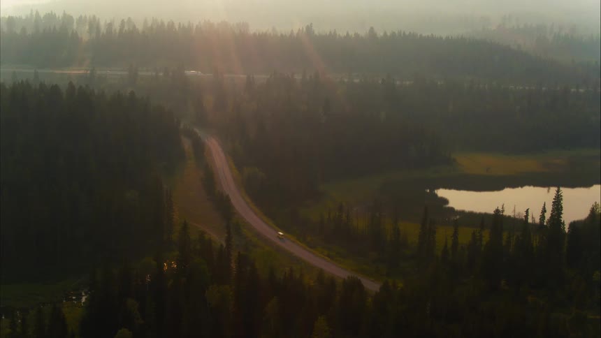 A lone car drives down a winding road through a vast, hazy forest landscape at golden hour