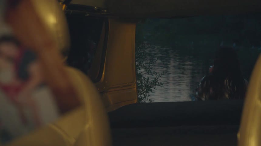 An old photo of a mother and child inside a car loses focus to reveal a solitary woman smoking by a dark lake
