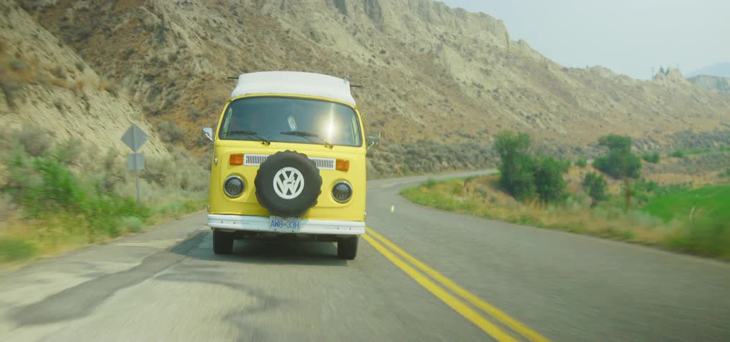 A classic yellow Volkswagen van enjoys a sun-drenched road trip along a winding country highway through the mountains