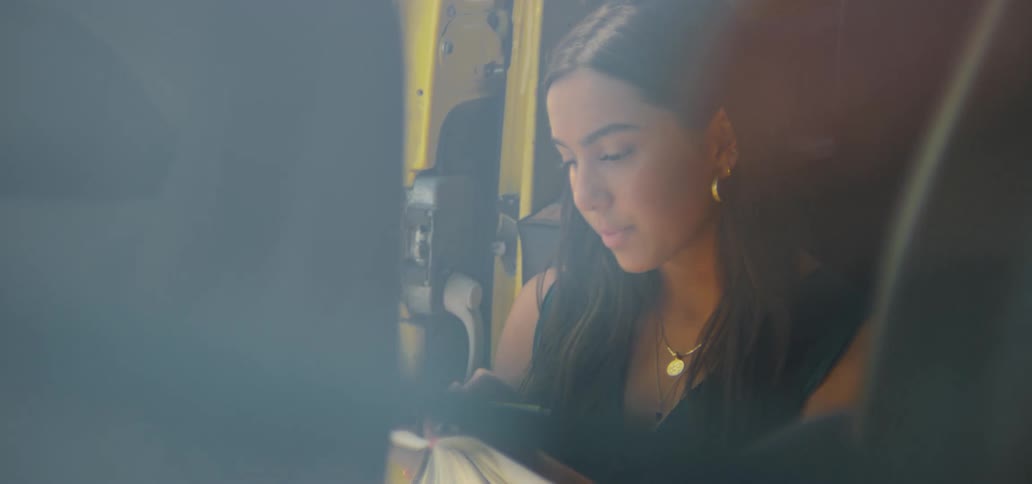 A pensive young woman journals her thoughts while sitting in the open door of her yellow vehicle during a road trip