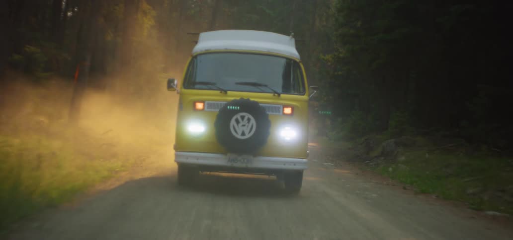 A classic yellow camper van kicks up dust as it drives down a forest road with its headlights on during a hazy golden sunset