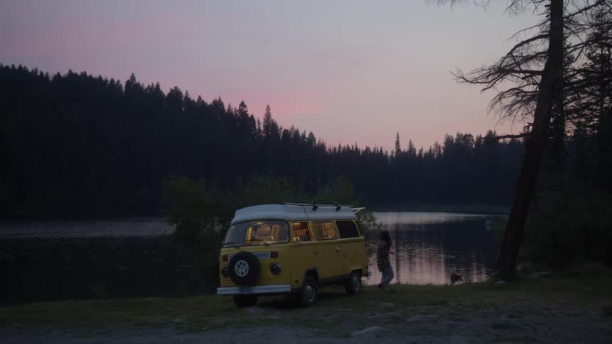 A woman and her dog get out of their cozy, lit-up vintage camper van to enjoy the peaceful lakeside view at dusk