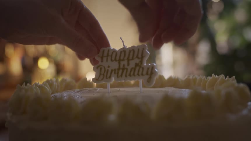 A close up of hands carefully placing a 'Happy Birthday' candle onto a freshly frosted cake in slow motion