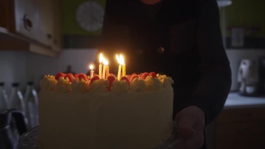 Slow motion close-up of a person carrying a birthday cake with lit candles and raspberries through a dimly lit room
