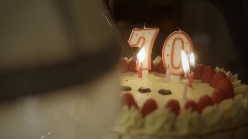 A person uses a lighter to ignite the number 70 candles on a decorated strawberry birthday cake in slow motion